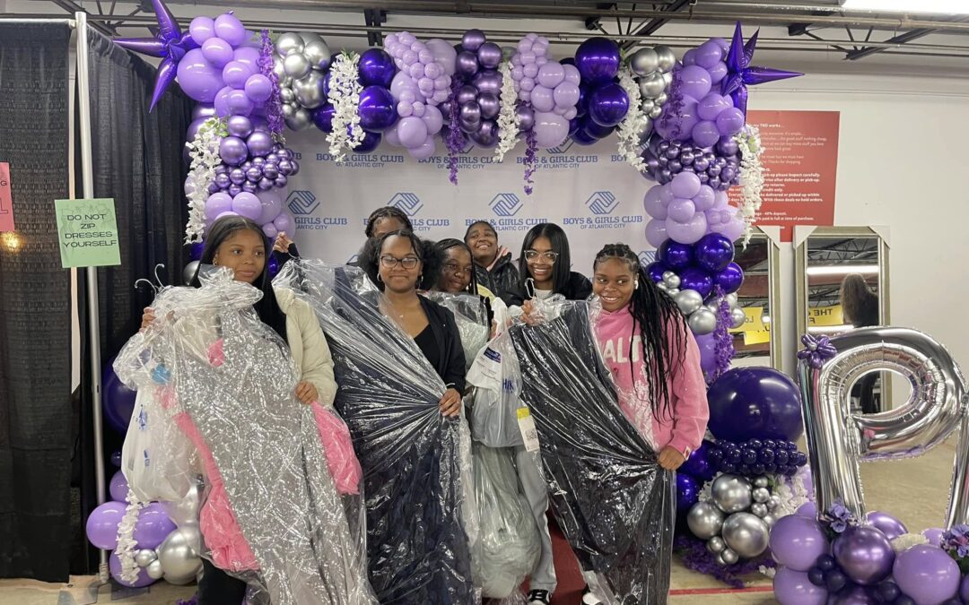 Girls in front of Project Prom backdrop, holding dresses